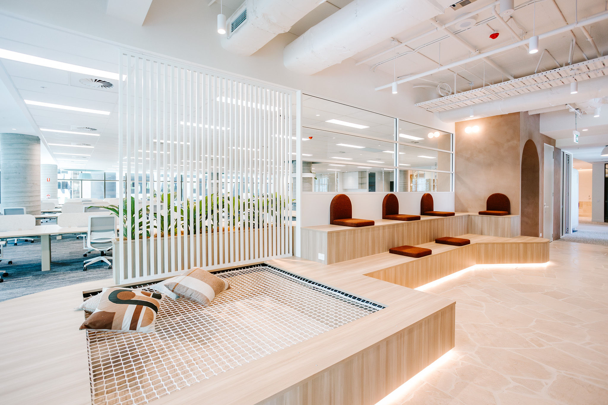 A modern office breakout zone featuring tiered timber seating with rust-toned cushions, a suspended hammock-style net lounge with patterned pillows, a white slatted partition with integrated greenery, and an open-plan workspace visible beyond, demonstrating TACTIC's Workplace Strategy approach to designing high-performing, adaptable environments.