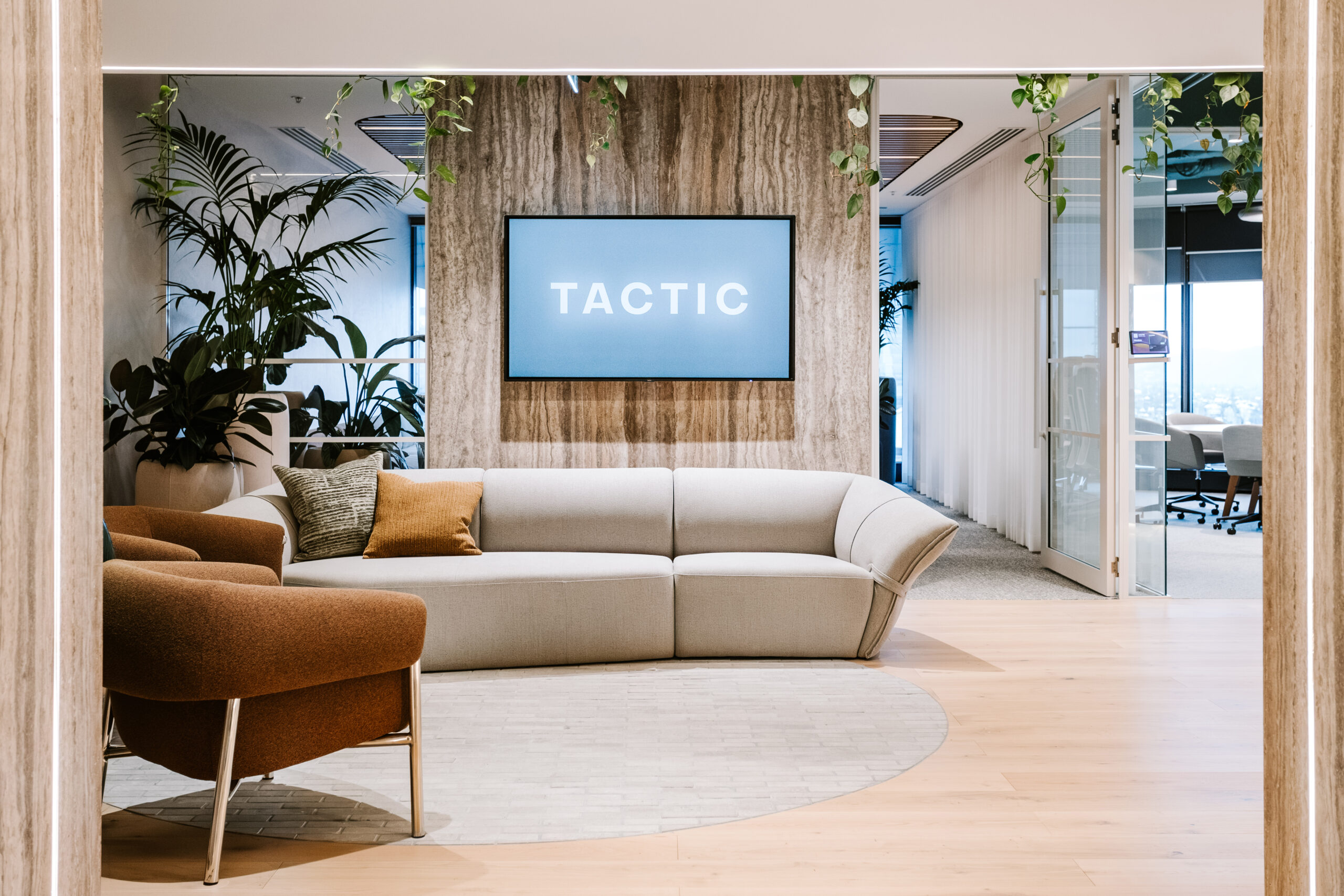 The TACTIC office reception and client lounge featuring a curved modular sofa, rust-toned velvet armchairs, integrated greenery, and a branded screen on a travertine feature wall, demonstrating strategy-led workplace design and fixed-price fitout delivery.